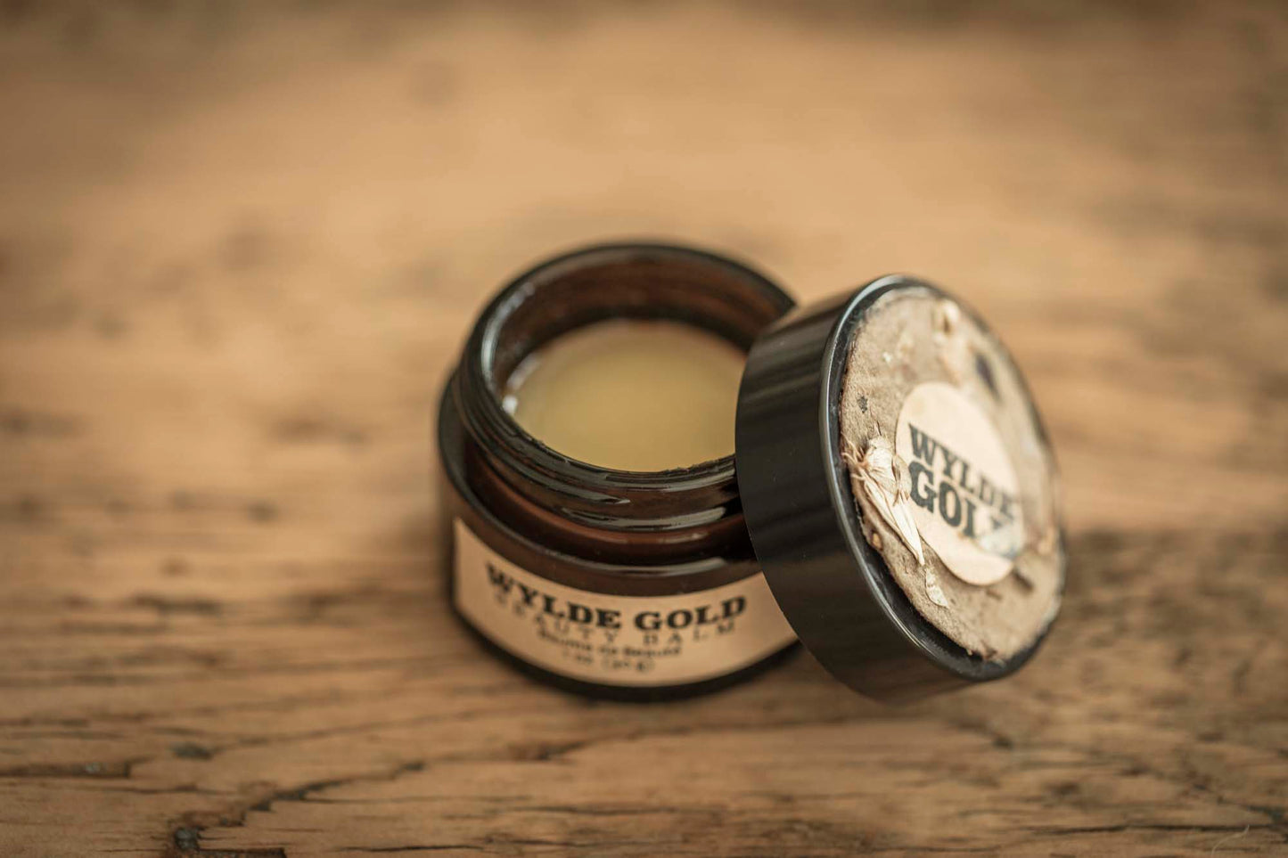 Open jar of Wylde Gold Beauty Balm with smooth golden balm surface, lid resting beside it on a wooden table.
