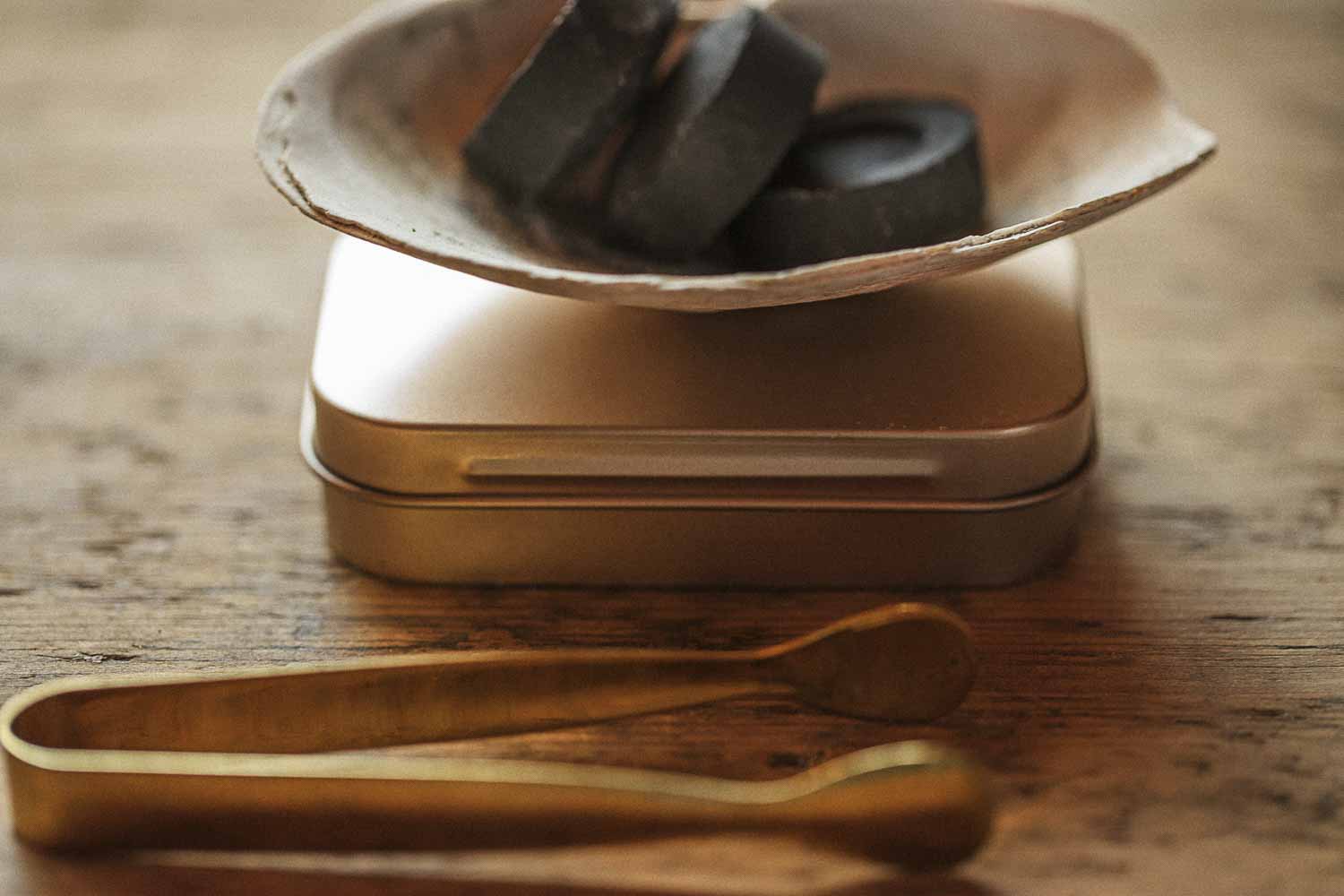 Shell burner with charcoal discs resting on a brass ritual tin, with brass tongs on a rustic wooden table.