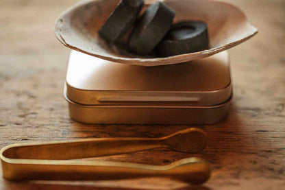 Shell burner with charcoal discs resting on a brass ritual tin, with brass tongs on a rustic wooden table.