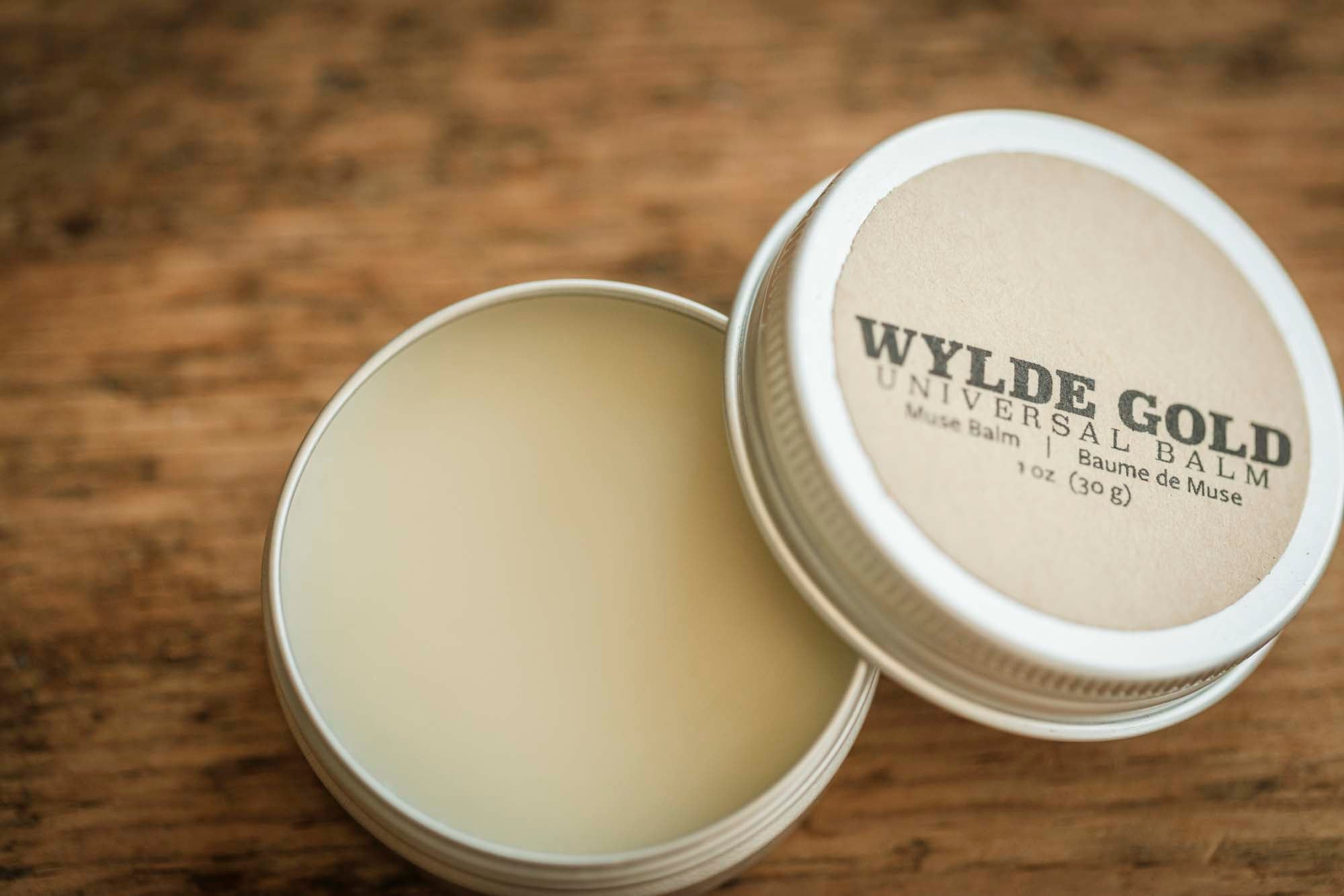 Open tin of Wylde Gold Universal Balm (Muse Balm) resting on a rustic wooden surface, showing smooth, creamy balm inside.