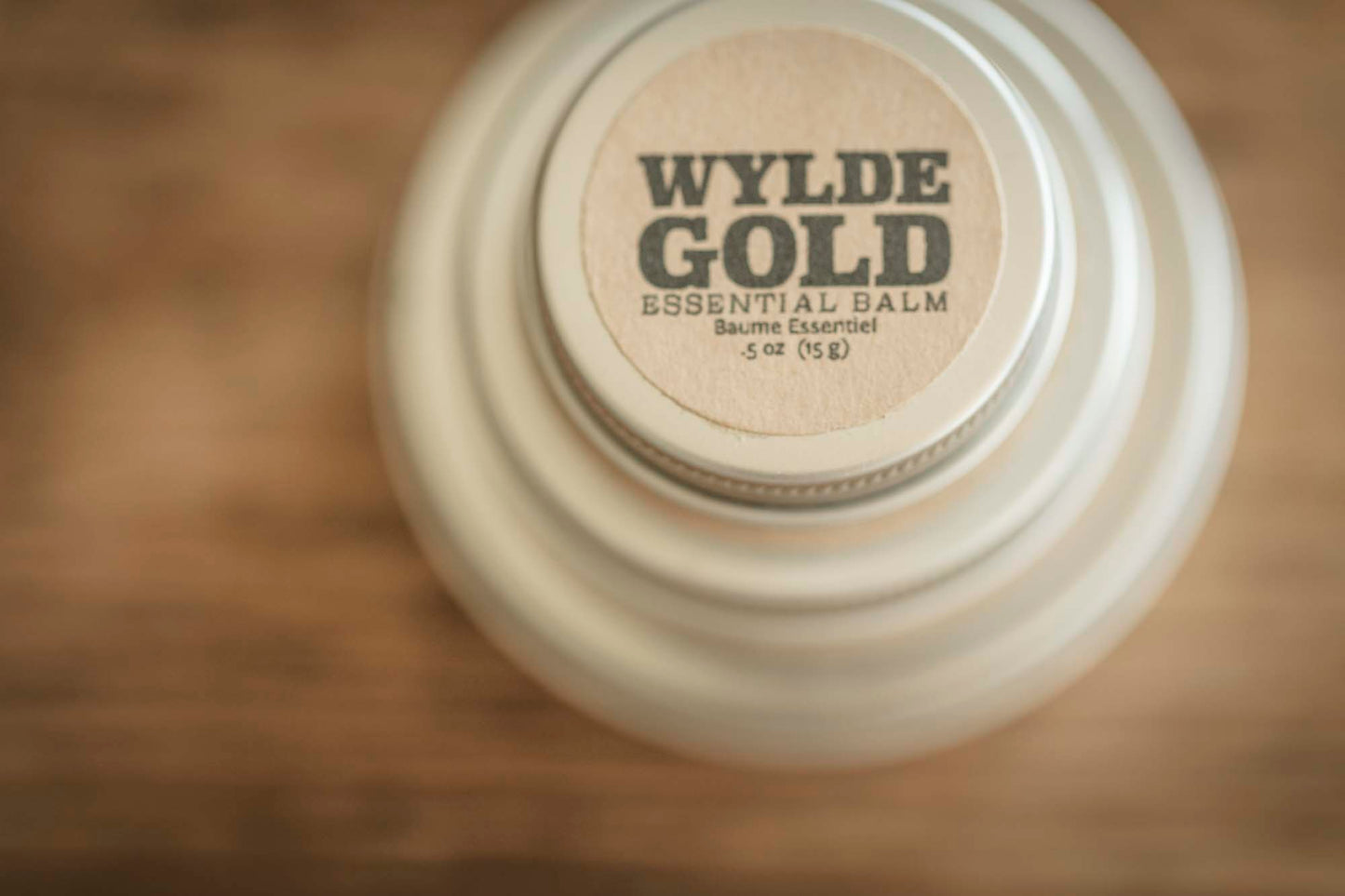 Top-down view of Essential Balm tin with centered WYLDE GOLD logo on a warm wood background.