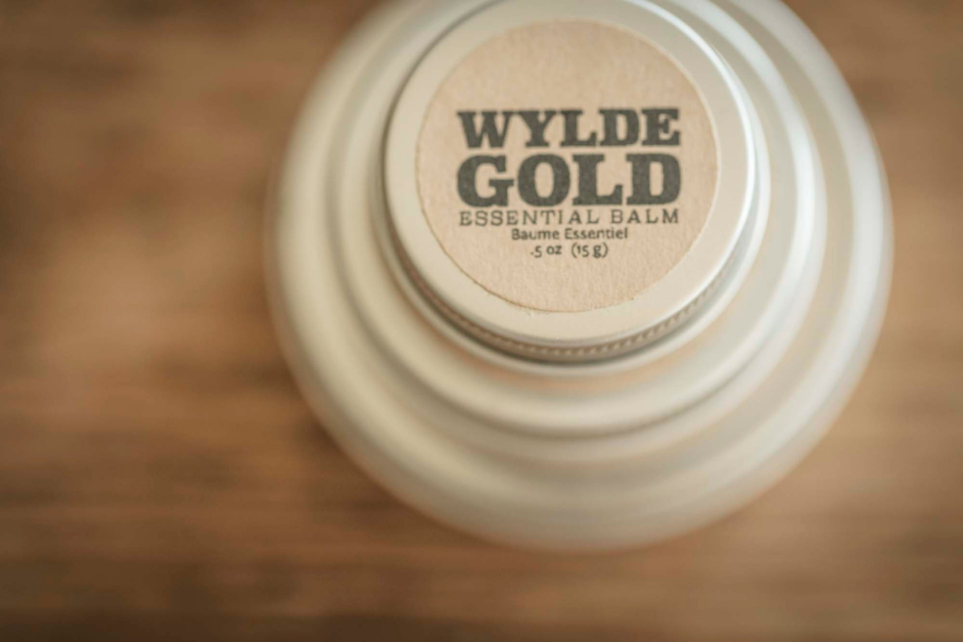 Top-down view of Essential Balm tin with centered WYLDE GOLD logo on a warm wood background.