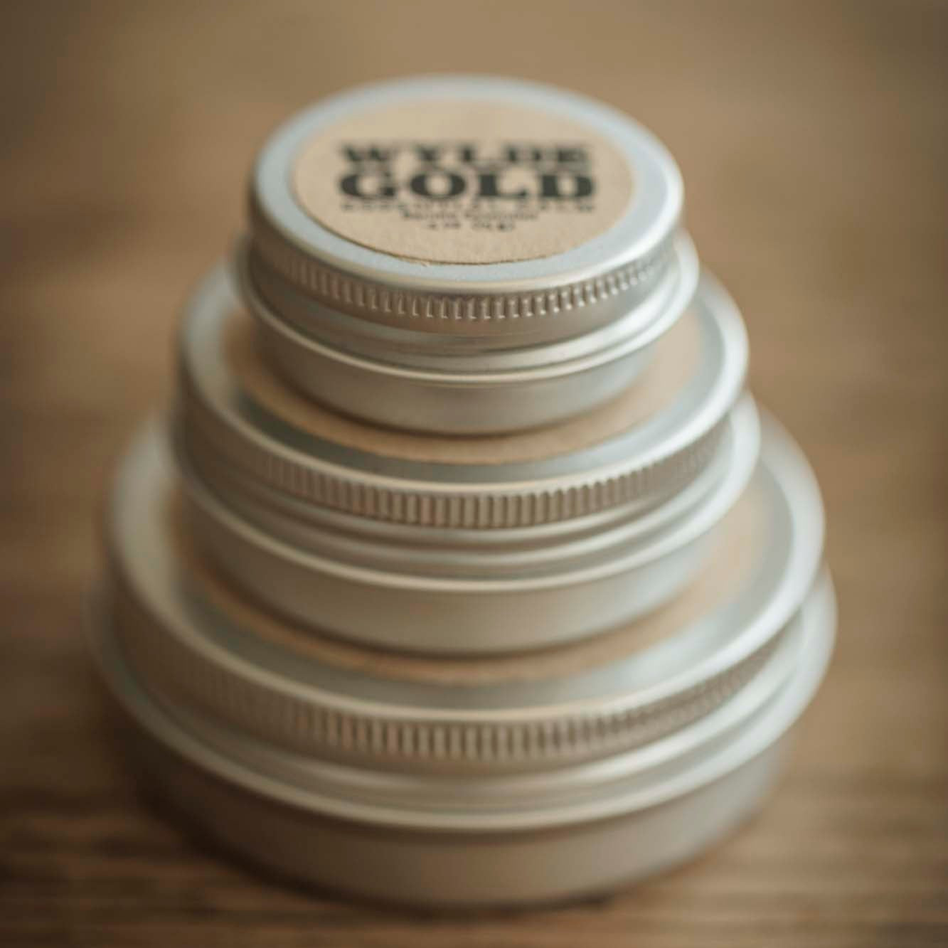 A stacked tower of Wylde Gold balm tins in descending size order, set on a warm wooden surface. The top tin is labeled with the WYLDE GOLD logo, creating a minimal, earthy, and elegant display.
