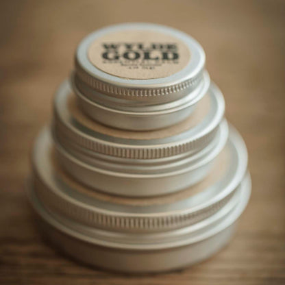 A stacked tower of Wylde Gold balm tins in descending size order, set on a warm wooden surface. The top tin is labeled with the WYLDE GOLD logo, creating a minimal, earthy, and elegant display.