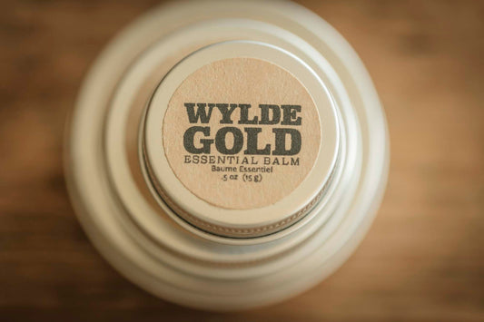 Essential Balm tin with WYLDE GOLD logo, photographed straight-on against a warm wooden background.