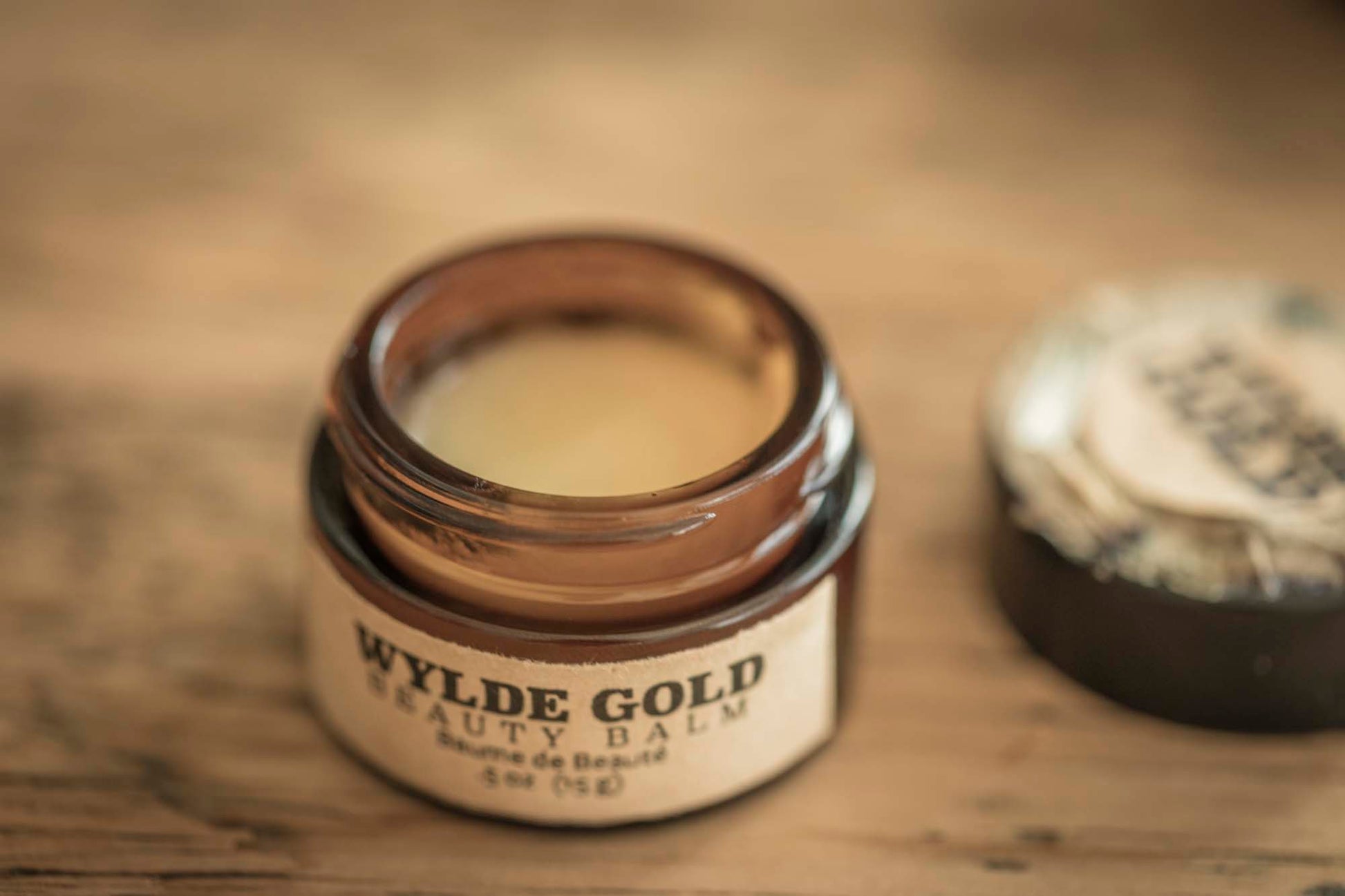 Open jar of Wylde Gold Beauty Balm with smooth golden balm surface, lid resting beside it on a wooden table.