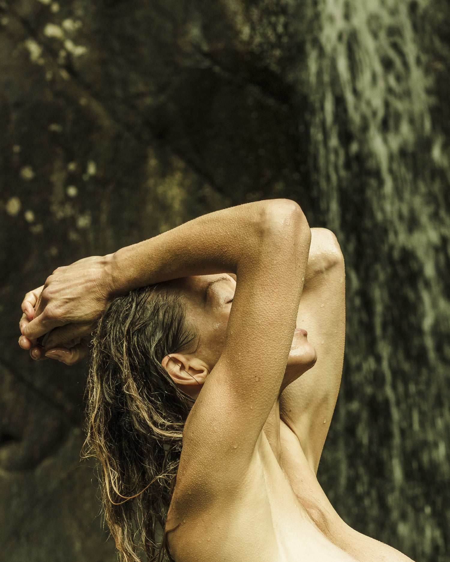 WYLDE GOLD | Woman standing topless in a waterfall, head tilted back, surrounded by trees in a raw, earthy moment of return